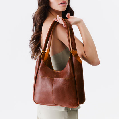 Nutmeg*Large | A model holding a u-shaped, flat-bottomed leather shoulder bag with two outside pockets