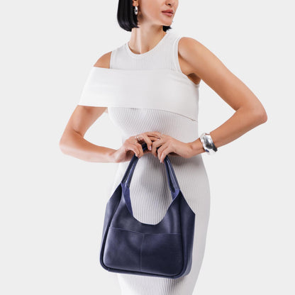 Cobalt*Medium | A model holding a u-shaped, flat-bottomed leather shoulder bag with two outside pockets